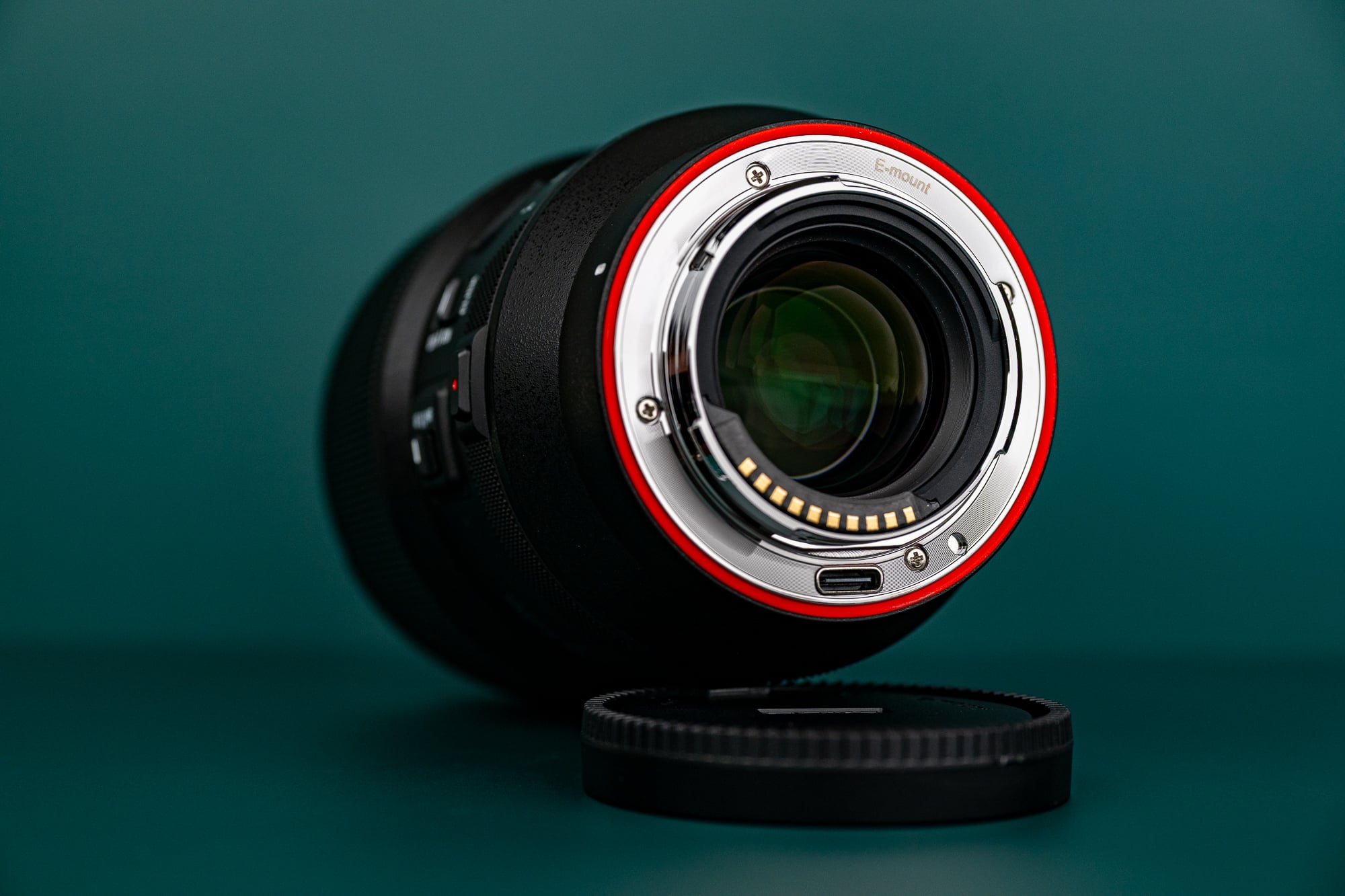 A camera lens positioned on its side with the metal lens mount facing forward, next to a detached lens cap, set against a dark green background.