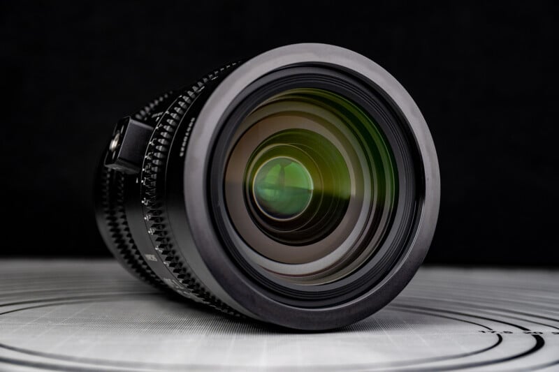 A close-up of a camera lens with visible glass elements and reflections, resting on a white surface with circular patterns, against a black background.