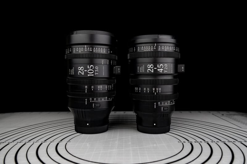 Two professional camera lenses with detailed markings stand side by side on a patterned surface against a black background. The lenses display focal lengths and aperture values.