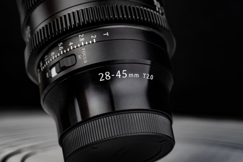Close-up of a black camera lens showing "28-45mm T2.0" and a focus adjustment switch, set against a blurred, dark background.