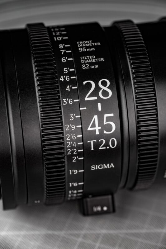 Close-up of a Sigma camera lens showing detailed focus and aperture markings, including "28-45 T2.0" and filter size "82mm" with textured grip rings visible.