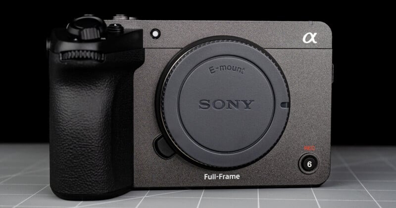 A close-up of a Sony Alpha full-frame mirrorless camera body with no lens attached, showing the E-mount and various control buttons on a gray grid-patterned surface.