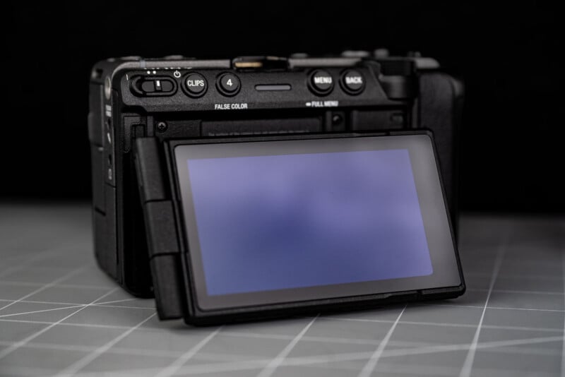 A digital camera with its rear LCD screen flipped out, facing forward. The camera sits on a gray surface with a grid pattern, and the background is black.