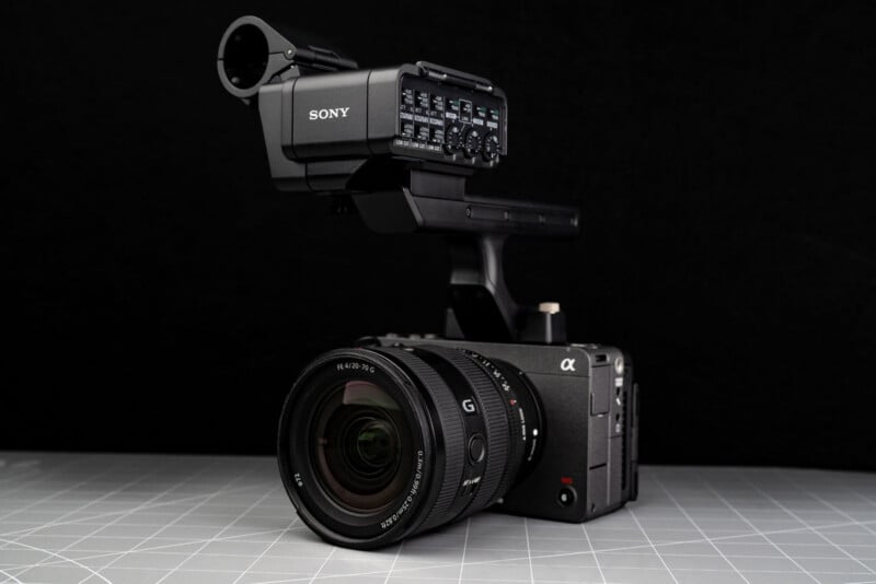 A professional Sony digital video camera with a large lens and external audio interface is placed on a gray grid-patterned surface against a black background.