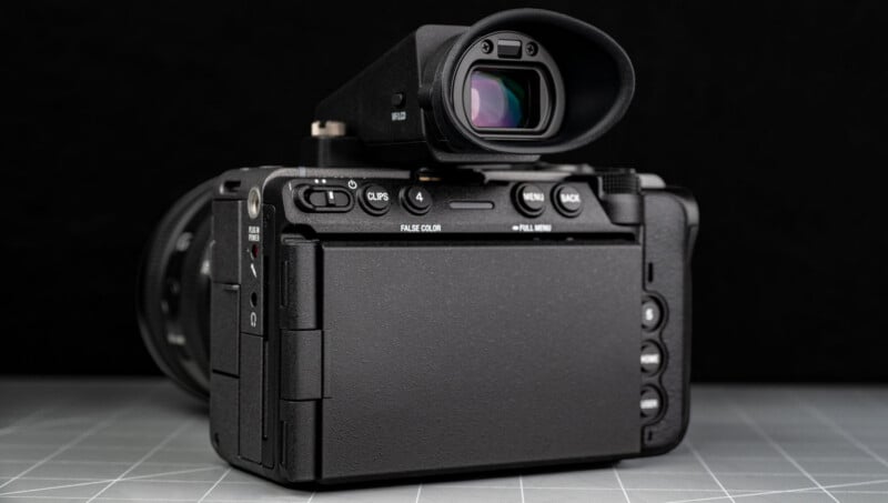 A digital camera with a closed flip-out screen is shown from the back, placed on a grid-patterned surface, with a black background.