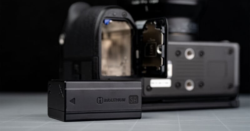 A camera with its battery compartment open is in the background, while a black InfoLITHIUM battery lies in the foreground on a flat surface.