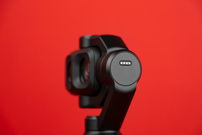 A black gimbal camera head with four gold electrical contact pins on its side, set against a solid red background.