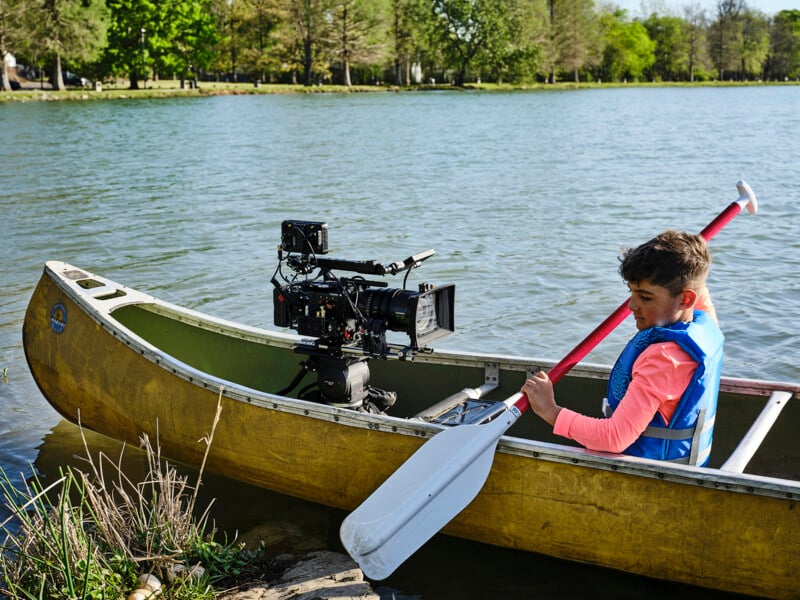 A boy in a blue life jacket sits in a yellow canoe on a lake, holding a paddle, with a large camera mounted in front of him. Trees and water are visible in the background.
