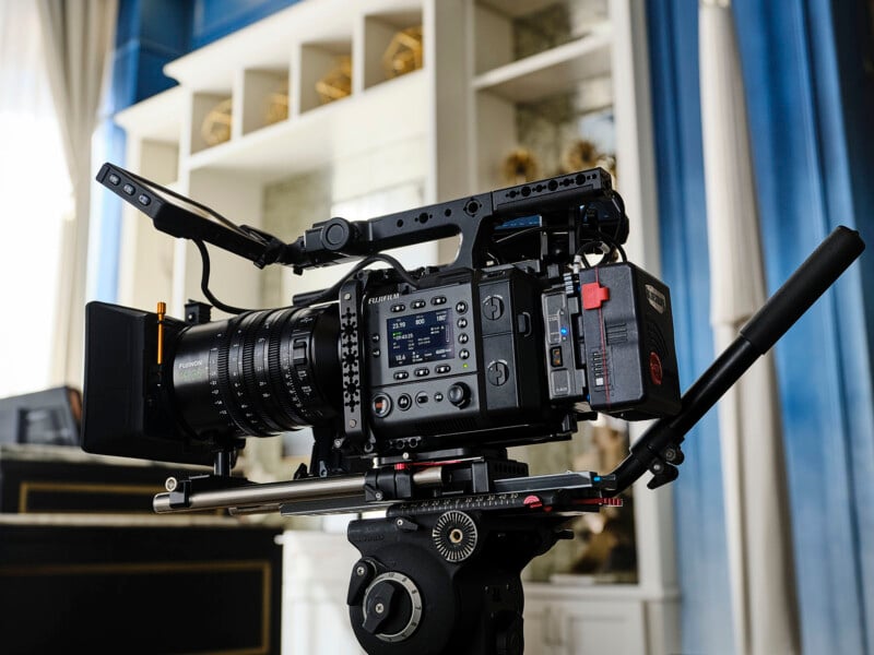 A professional digital cinema camera with various attachments and a large lens is mounted on a tripod indoors, in front of blue and white shelves.