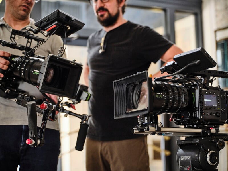 Two people stand indoors, holding professional video cameras with large lenses and mounted screens. A third camera is positioned on a tripod in the foreground, focused and ready for filming.