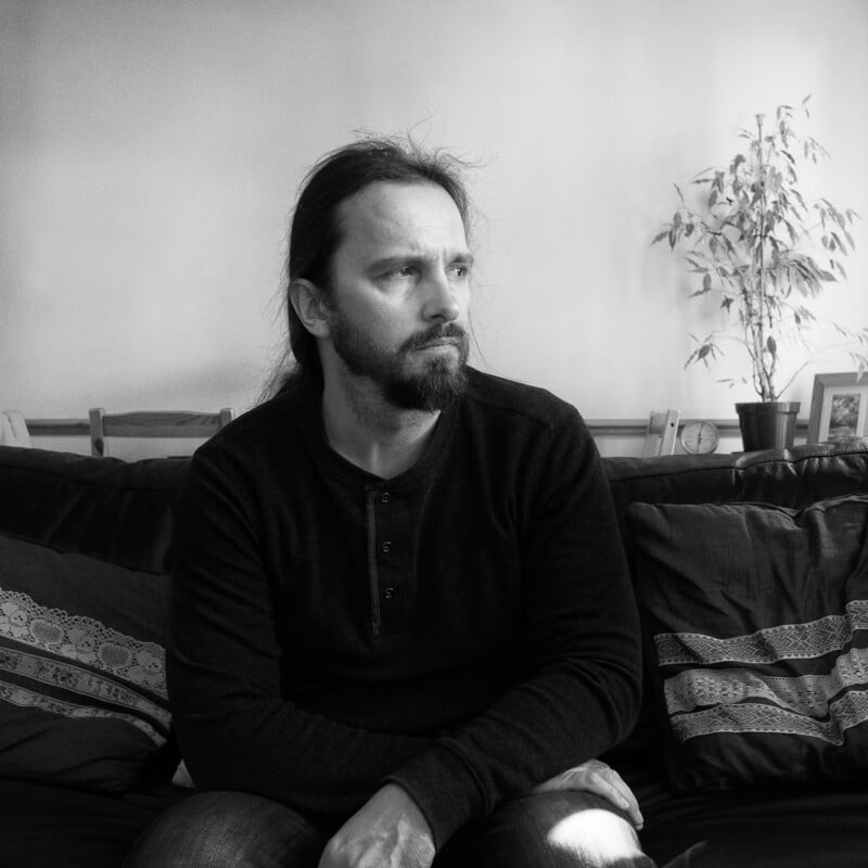 A man with long hair and a beard sits on a couch, looking to the side. He wears a dark shirt and is in a softly lit room with a plant and framed photo in the background.