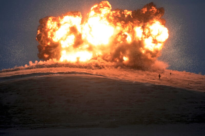 A large fiery explosion erupts on a hilltop under a dark sky, with thick smoke and bright flames spreading outward and two small human figures visible in the distance.