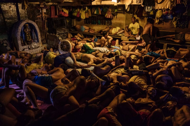 Dozens of men sleep closely packed together on the floor of a dimly lit, overcrowded room, with clothing and personal items hanging above them.