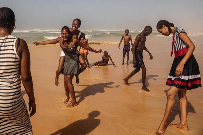A group of people play and laugh together on a sandy beach, with some standing in the shallow water and one person giving another a piggyback ride. The ocean waves are visible in the background.