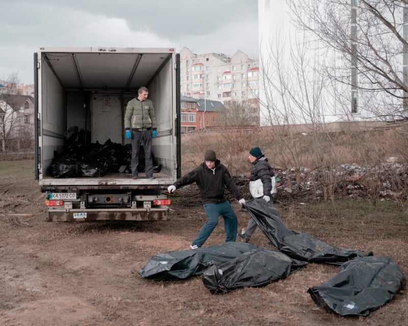Two people in winter clothing move black body bags from the ground toward a truck, where another person stands among more body bags. Urban buildings and bare trees are visible in the background.