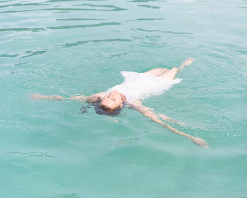 A person in a white dress floats on their back in clear turquoise water, arms and legs outstretched, appearing relaxed and peaceful.
