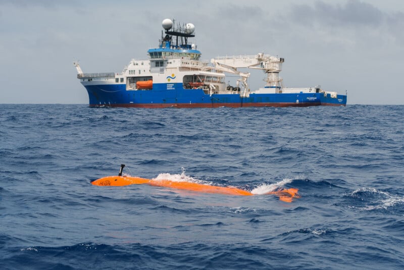 A bright orange underwater drone floats on the ocean surface, with a large blue and white research vessel in the background under a cloudy sky.