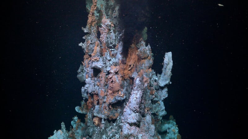 A tall underwater hydrothermal vent emits dark smoke from its top, with rugged, mineral-coated surfaces and deep-sea particles floating in the surrounding dark water.