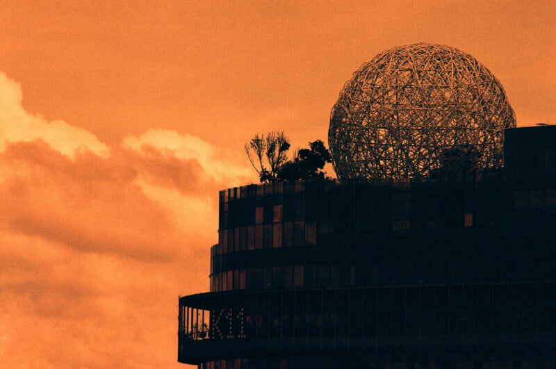 A modern building with a large wireframe sphere and trees on its rooftop is silhouetted against an orange sky with clouds.