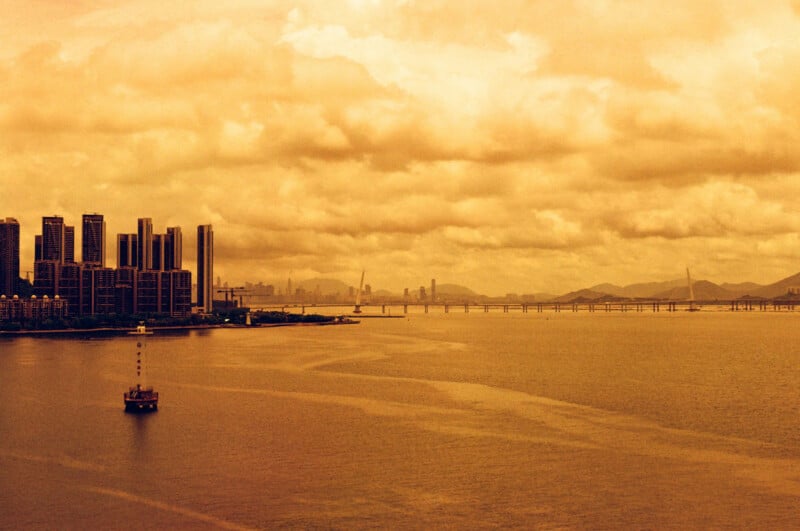 A city skyline with tall buildings sits by a wide body of water. A boat floats in the foreground, and a long bridge stretches into the distance under a cloudy, golden-hued sky. Distant mountains are visible.