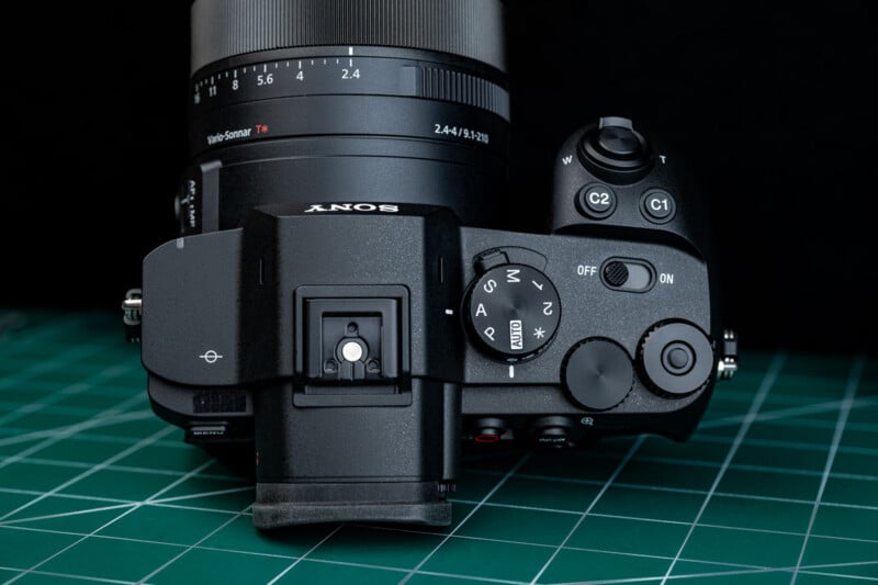 Top view of a Sony digital camera with attached lens, placed on a green cutting mat. Camera dials, buttons, and controls are clearly visible, including the mode dial and on/off switch.