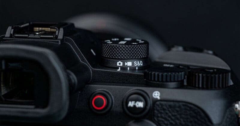 Close-up of a black camera showing the mode dial with "S&Q" selected, a red button, and various control dials against a dark background.