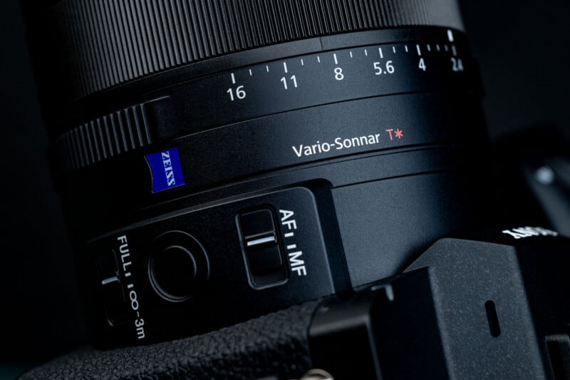 Close-up of a camera lens showing "Vario-Sonnar T*" and "Zeiss" labels, with focus and AF/MF switches visible. The textured lens ring and part of the camera body are in view.
