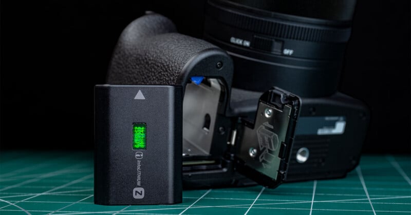 A camera sits on a green cutting mat with its battery compartment open and a camera battery partially removed, showing the battery status indicator glowing green.