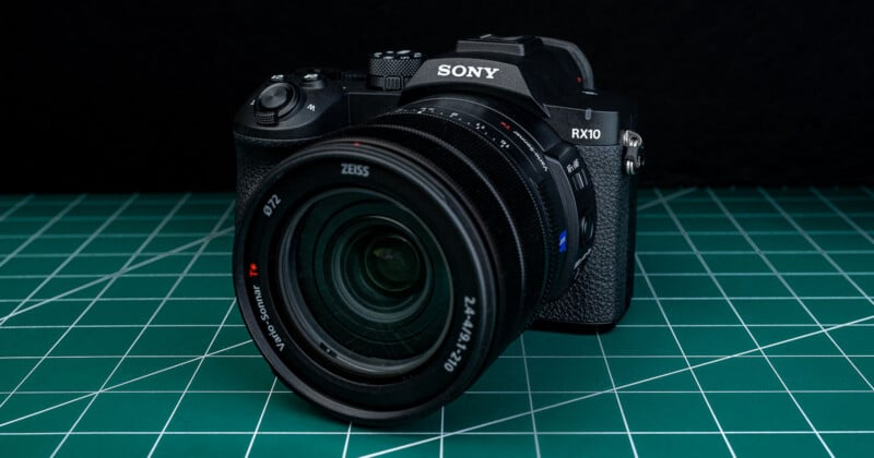 A Sony RX10 camera with a Zeiss lens is placed on a green cutting mat with a grid pattern. The background is black, highlighting the camera's details and texture.