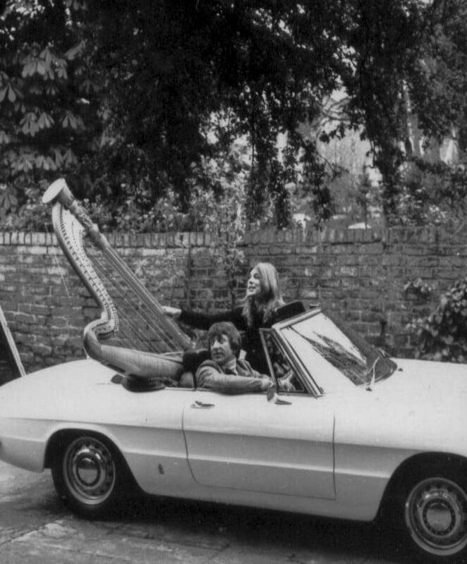 A man and a woman sit in a white convertible car with a large harp resting in the back seat.