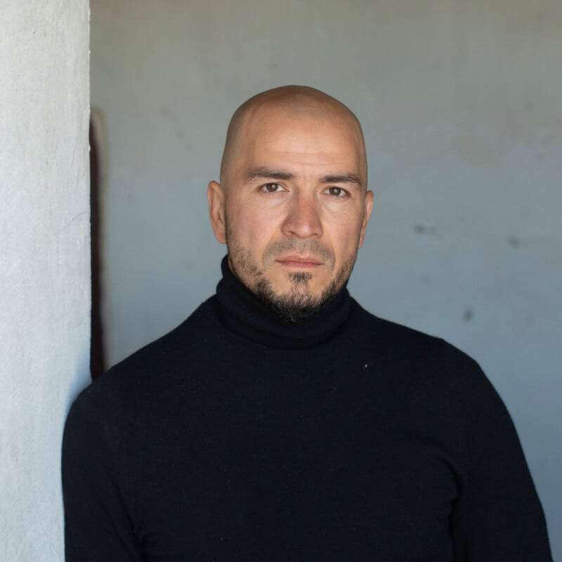 A man with a shaved head and trimmed beard wears a black turtleneck sweater, standing against a plain, light-colored wall, looking directly at the camera with a neutral expression.