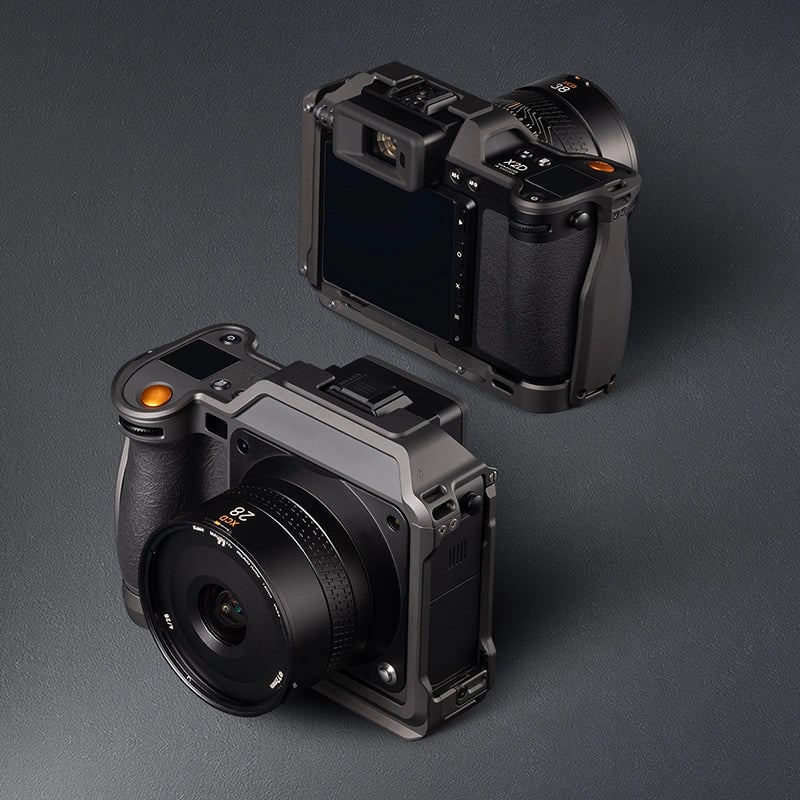Two modern digital cameras are displayed on a dark surface, one showing the lens and front controls, the other showing the rear touchscreen and viewfinder. Both are sleek, black, and compact, with textured grips and orange shutter buttons.