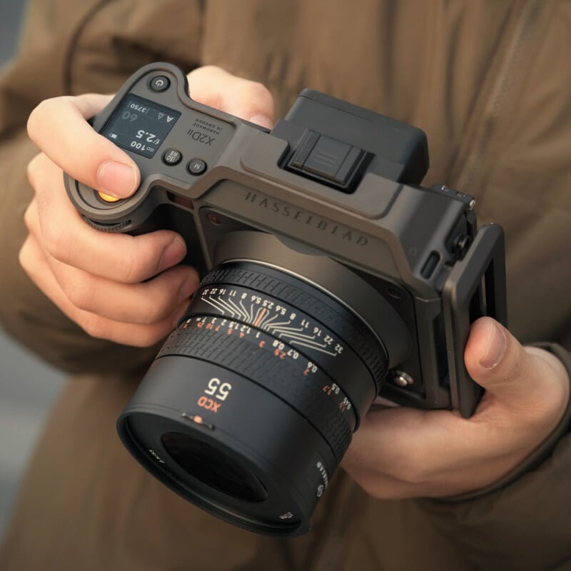 Close-up of hands holding a Hasselblad medium format camera, focusing on the lens and controls. The person is wearing a brown jacket.