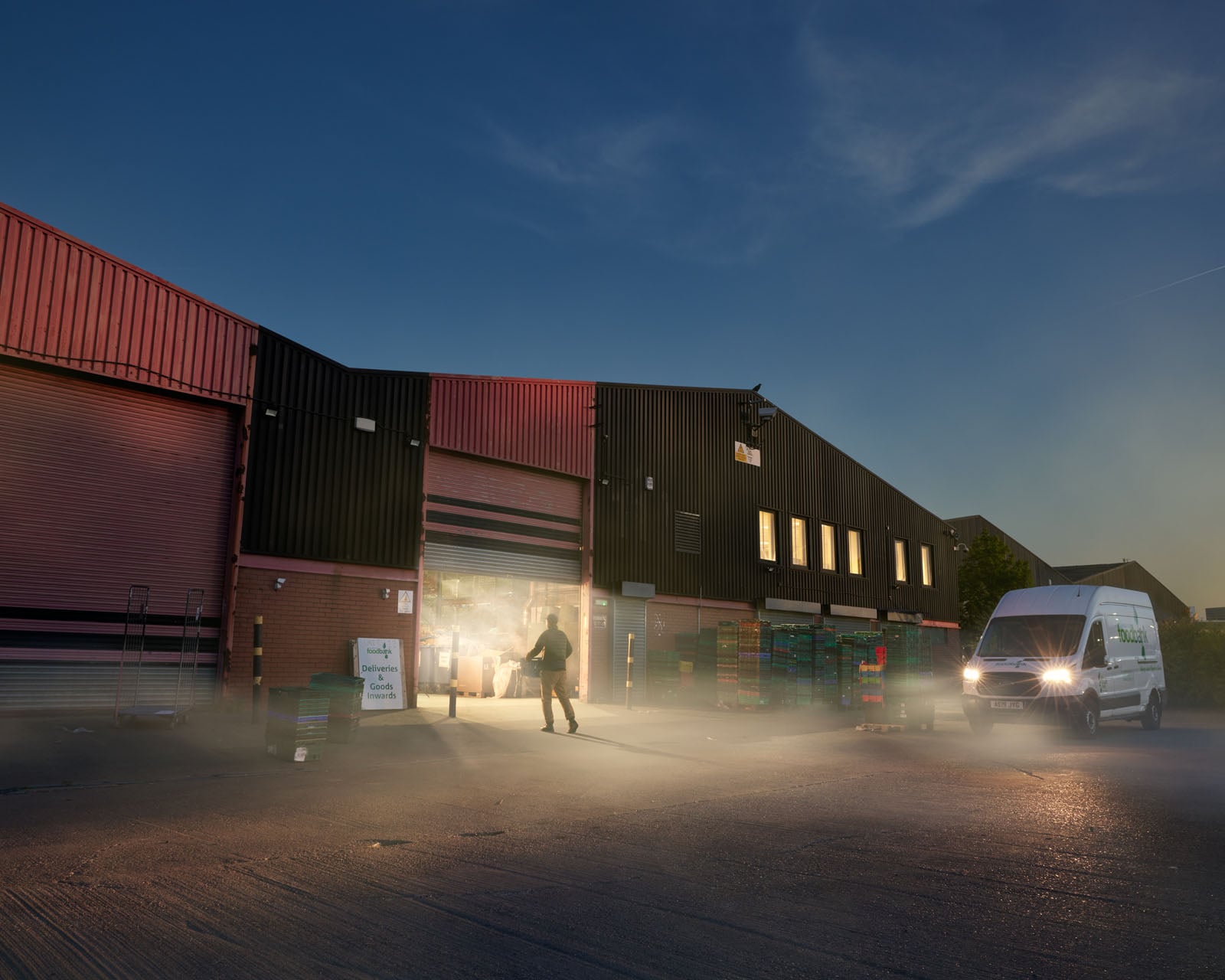 A person walks toward the open bay of a warehouse building at dusk, with a white delivery van parked nearby in the haze.
