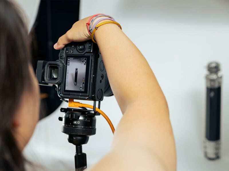 A person takes a photo of a cylindrical object with a digital camera on a tripod. The camera screen shows the object in focus against a plain background.