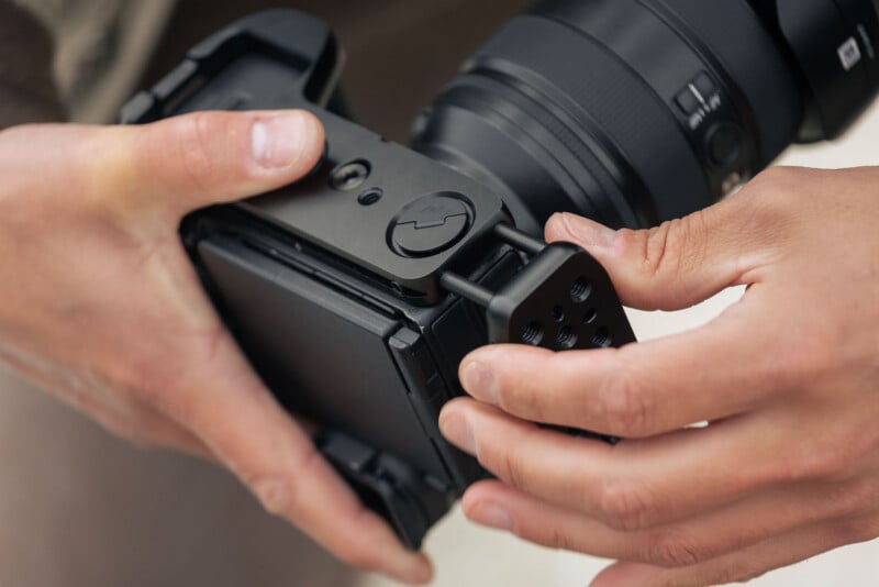 Close-up of a person’s hands attaching a black camera accessory, possibly a mounting bracket or tripod plate, to the bottom of a digital camera with a large lens.