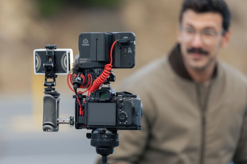 A camera with multiple attachments is set up on a tripod, focused on a man in a tan jacket with a mustache, who is blurred in the background.