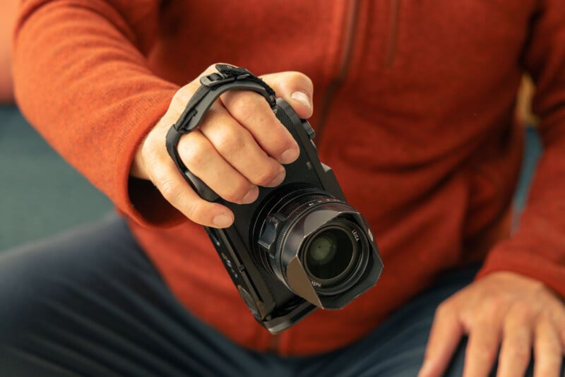 A person wearing an orange jacket holds a black camera with a strap, gripping it securely in their right hand while sitting down.