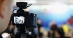 A close-up of a digital camera on a tripod filming a group of people in a brightly lit, blurred background setting, possibly during an interview or discussion.