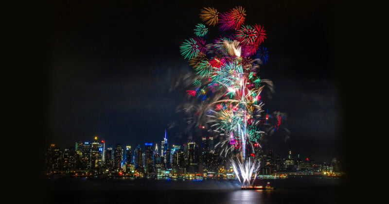 Colorful fireworks burst over a city skyline at night, reflecting on the water below. Bright city lights illuminate the buildings, creating a festive and vibrant atmosphere.