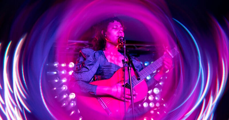 A musician plays guitar and sings into a microphone on stage, surrounded by vibrant pink and purple swirling light effects.