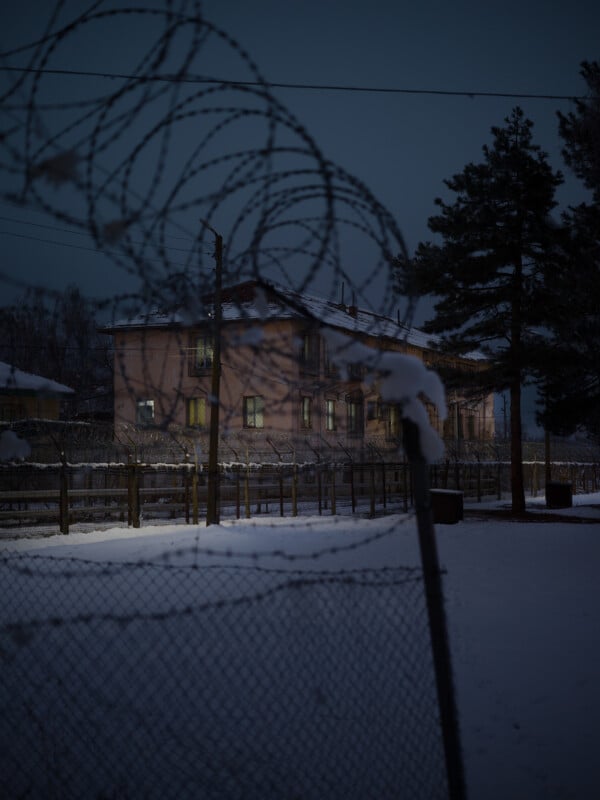 A snow-covered building is seen through barbed wire and a chain-link fence at dusk. Dim lights glow from the windows, and tall pine trees stand nearby. The scene feels cold and isolated.