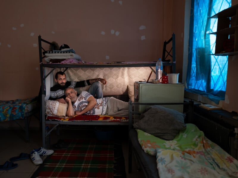 A man and woman lie together on a lower bunk bed in a modest room with peach walls, blue curtains, and sparse furniture, illuminated softly by a lamp. Personal belongings are visible on the beds and nearby surfaces.