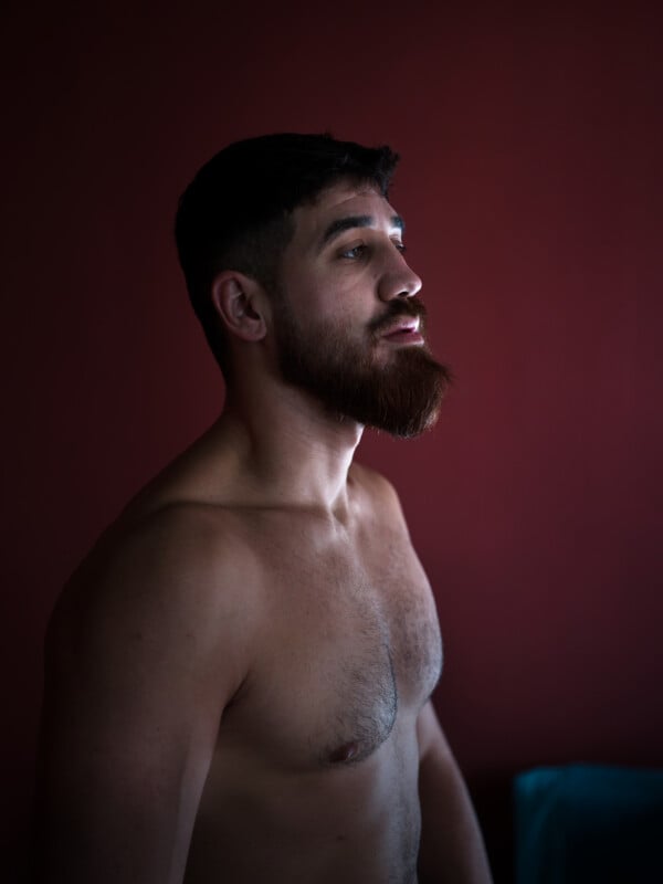 Shirtless man with a beard and short hair stands in profile against a dark red background, looking slightly upward.