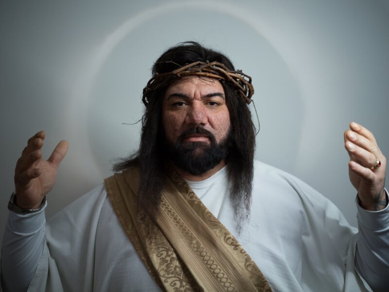 A person dressed in biblical-style robes and a crown of thorns raises both hands, with a halo-like circle behind their head, evoking a traditional depiction of Jesus.