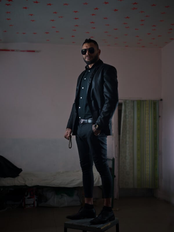 A man in dark sunglasses, black suit, and black shirt stands confidently on a stool in a dimly lit room with a patterned ceiling and curtain, holding prayer beads in one hand.