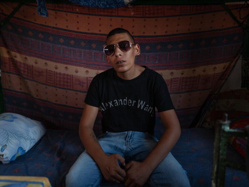 A young man wearing sunglasses and a black Alexander Wang t-shirt sits on a bed with a patterned blanket hanging behind him, hands resting on his lap, in a dimly lit room.