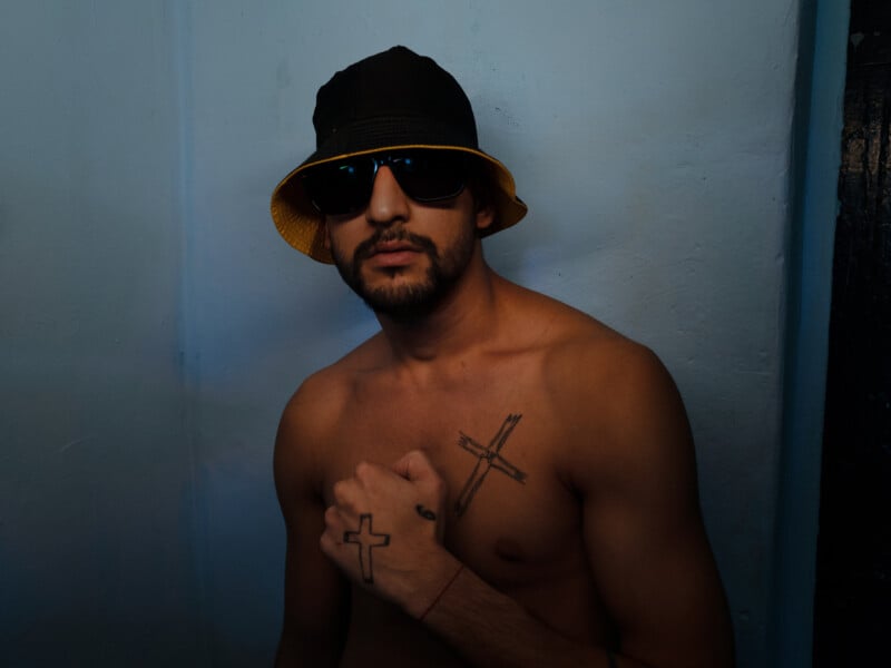 A shirtless man wearing sunglasses and a black bucket hat stands against a blue wall, showing tattoos of crosses on his chest and hand.