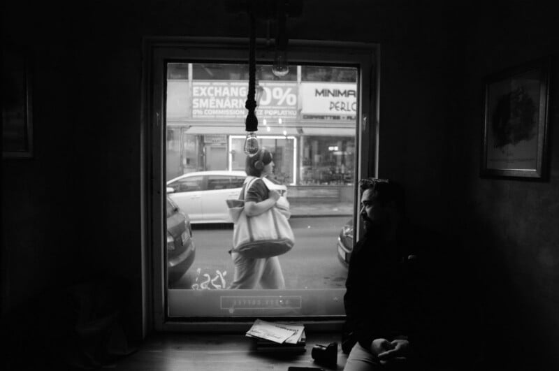 A person sits in a dark room near a window, looking out. Outside, a person with bags walks past parked cars and signs on the street. Light from the window highlights their silhouette.