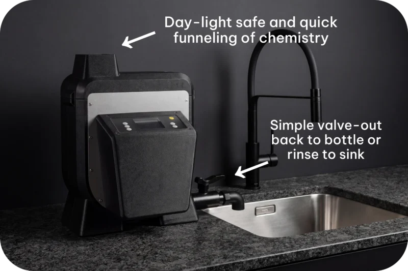 A black film processing machine sits on a granite countertop next to a kitchen sink, with text labels pointing to its top funnel and side drain valve.